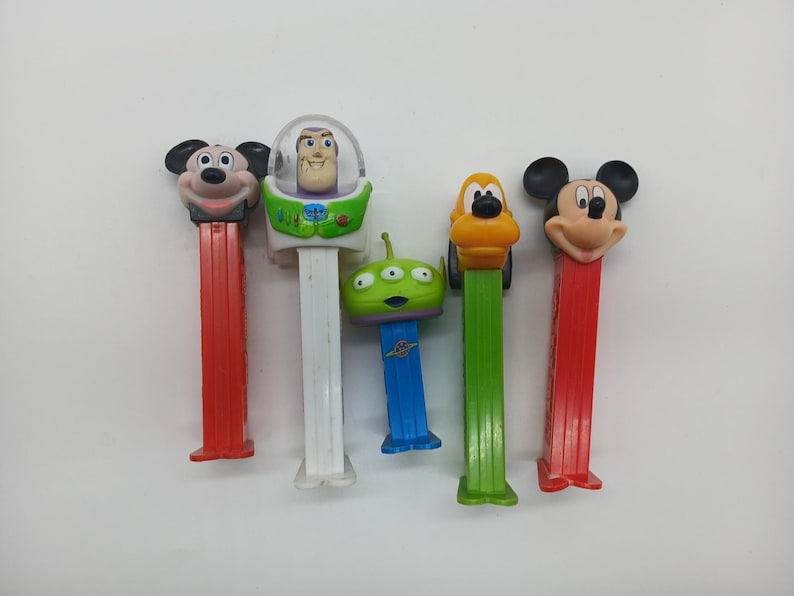 May include: A collection of five PEZ dispensers featuring popular cartoon characters. The dispensers include Mickey Mouse, Buzz Lightyear, a Little Green Alien, and Pluto. Each dispenser has a colorful design and a rectangular base.