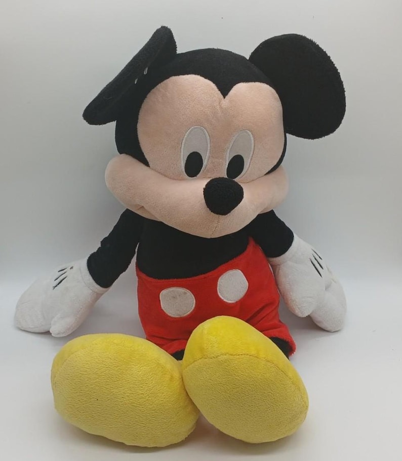 以下が含まれることがあります： ミッキーマウスのぬいぐるみ。象徴的なキャラクターは、黒い頭と耳、ピンクの顔、黒い鼻をしています。白いボタンが付いた赤いショーツ、黒い腕、白い手袋、黄色の靴を履いています。おもちゃは白い背景に置かれています。