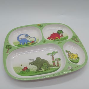 May include: A green divided plate with four sections, featuring cartoon dinosaurs. The plate has a light green border and white interior sections. Dinosaurs include a blue Brontosaurus, a red Stegosaurus, a green Triceratops, and a yellow Mammoth.