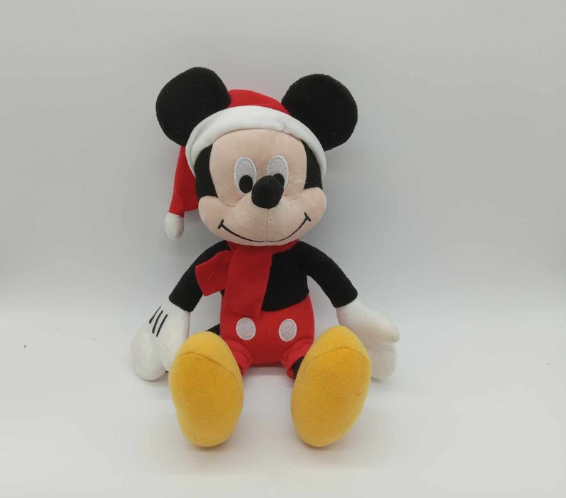May include: A plush Mickey Mouse toy wearing a Santa hat and red scarf. The toy has black ears, a black body, red shorts, and yellow feet. The toy is sitting with its arms and legs extended.