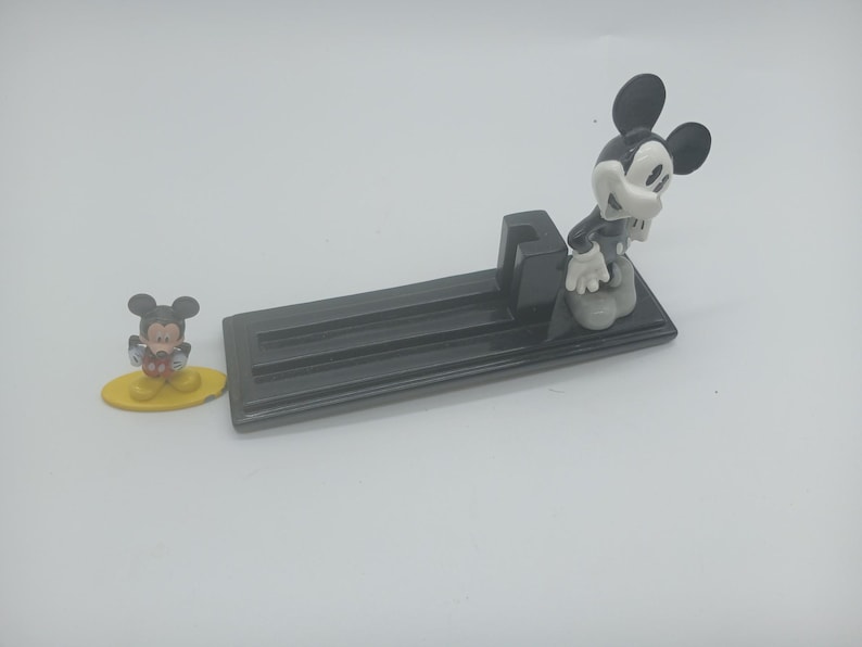 May include: A black rectangular ashtray with three grooves, featuring a Mickey Mouse figurine. A smaller Mickey Mouse figurine on a yellow base is also present. The figurines are in classic black, white, and yellow colors.