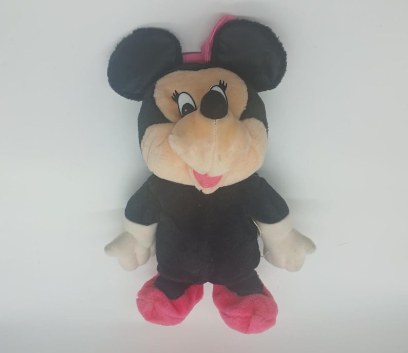 K&ouml;nnte beinhalten: Ein Pl&uuml;sch Minnie Mouse-Puppe mit schwarzen Ohren, einem schwarzen K&ouml;rper und einem beigen Gesicht. Die Puppe hat eine rosa Schleife, rosa Schuhe und wei&szlig;e Handschuhe. Beliebte Disney-Figur.