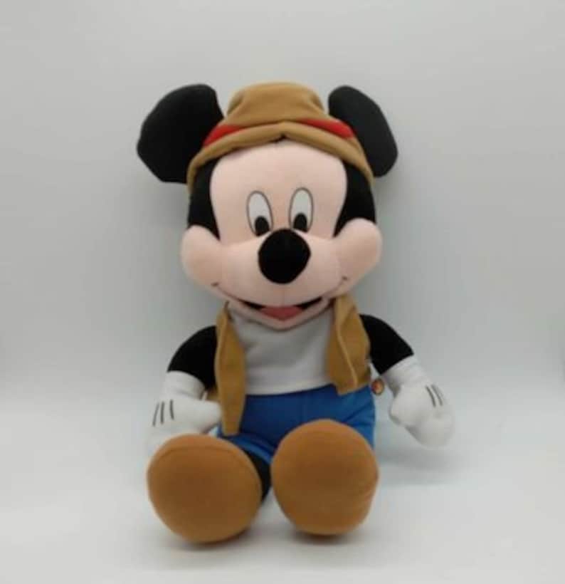 May include: A plush Mickey Mouse toy wearing a tan safari hat with a red band, a tan vest, blue shorts, and brown shoes. The toy has black ears, a black nose, and white gloves with three black lines.