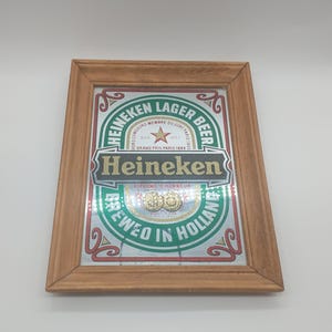 May include: A framed Heineken Lager Beer sign. The sign features a green and white design with the Heineken logo in gold and black. The frame is made of light brown wood.
