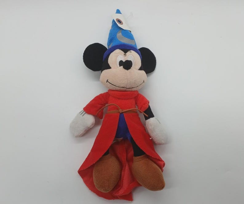 May include: A plush Mickey Mouse dressed as a sorcerer, wearing a blue hat with a crescent moon, a red robe, and brown shoes. The stuffed animal has black ears, a tan face, and white gloves. A classic Disney character.