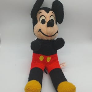 May include: Vintage Mickey Mouse plush toy. The plush features a black body, ears, and arms, with a tan face, red shorts, and yellow feet. The toy has a smiling expression and is in a sitting position. A Walt Disney tag is attached.
