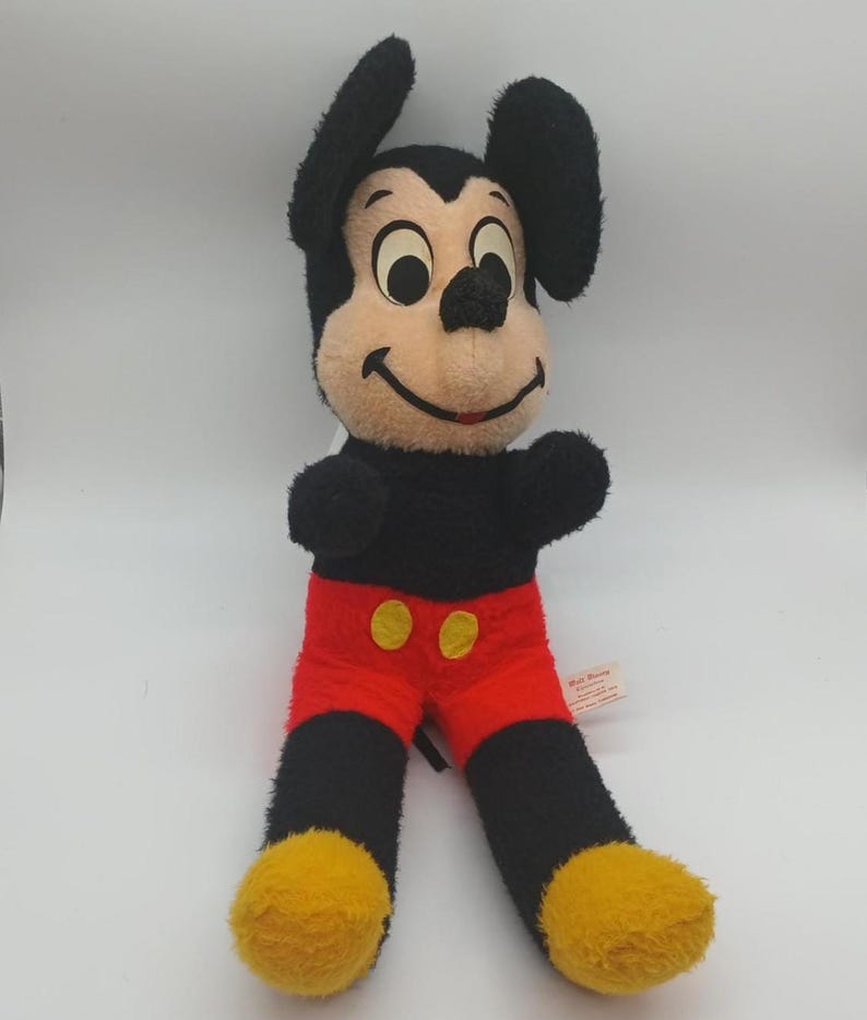 May include: Vintage Mickey Mouse plush toy. The plush features a black body, ears, and arms, with a tan face, red shorts, and yellow feet. The toy has a smiling expression and is in a sitting position. A Walt Disney tag is attached.