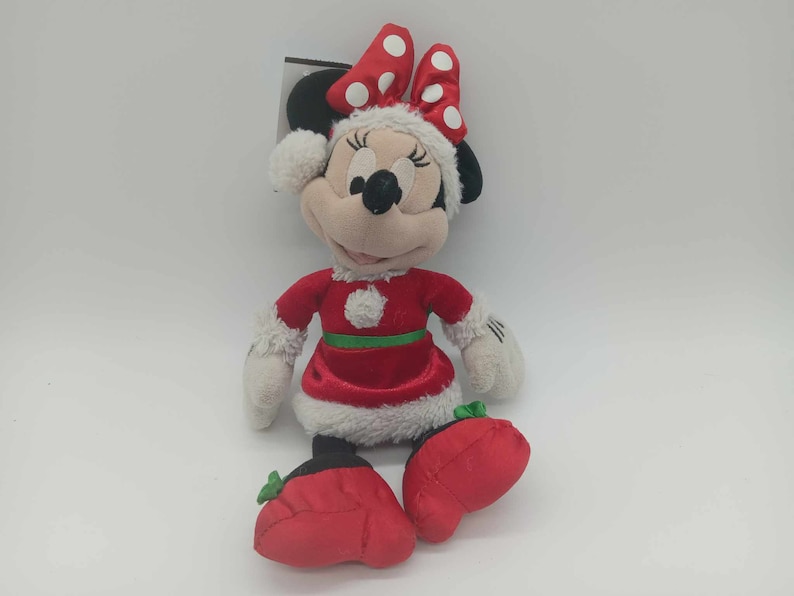 May include: A Minnie Mouse soft toy dressed in a Christmas outfit. The toy has a red dress with white trim, a green belt, and red shoes. Minnie's bow is red with white spots. The plush has black ears and a smiling face.
