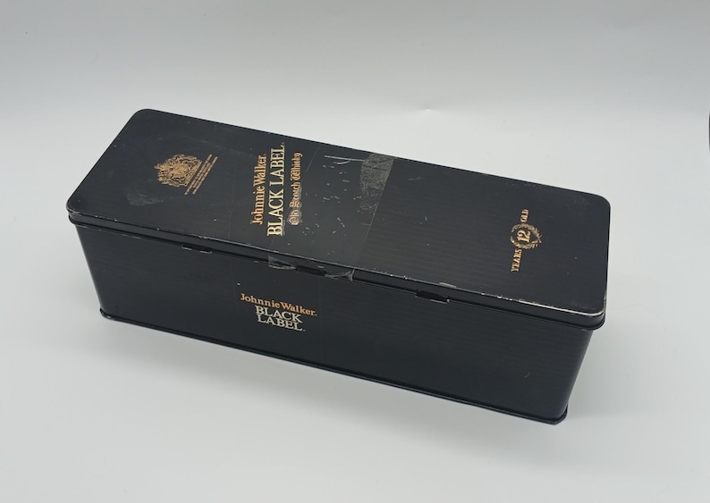 May include: A black rectangular box with gold lettering and a gold emblem. The text on the box reads "Johnnie Walker Black Label Scotch Whisky" and "Years 12 Old". The box is slightly worn.