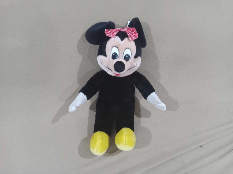 May include: Black plush toy of Minnie Mouse with a red bow and white polka dots. She has yellow shoes and a black nose.