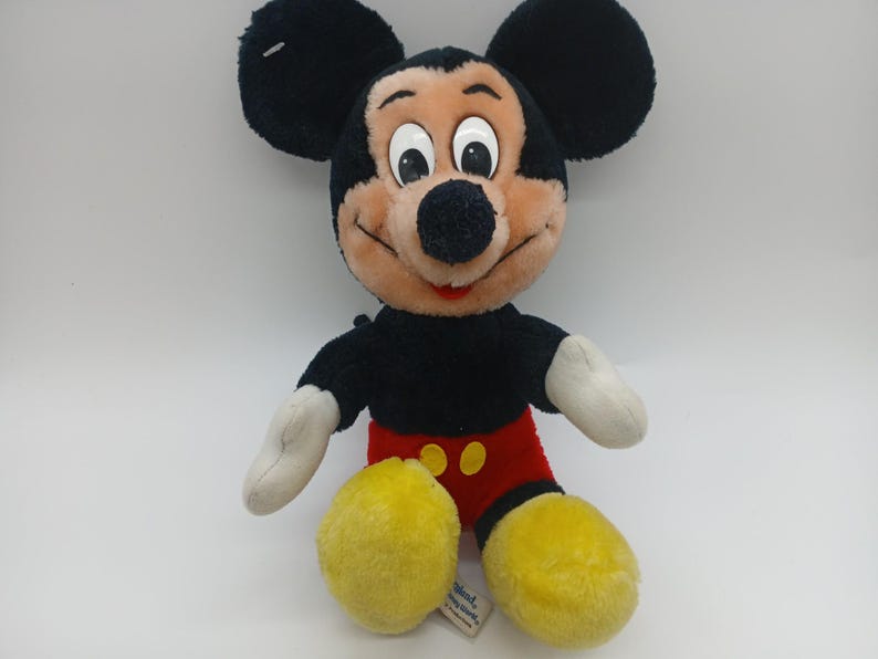 May include: A plush Mickey Mouse toy with black fur, large black ears, and a tan face. He wears red shorts with yellow buttons, yellow shoes, and white gloves. The toy is sitting on a white surface.