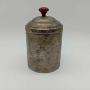 May include: A vintage silver-toned canister with a domed lid and a red knob. The canister has a cylindrical shape and shows signs of wear, with a mottled surface. The lid fits snugly on top.