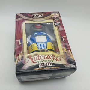 May include: A blue and yellow M&M's Nutcracker candy dispenser in its original packaging. The dispenser is shaped like an M&M's character wearing a Nutcracker costume. The packaging includes the text "Nutcracker Sweet M&M's Chocolate Candy Dispenser".