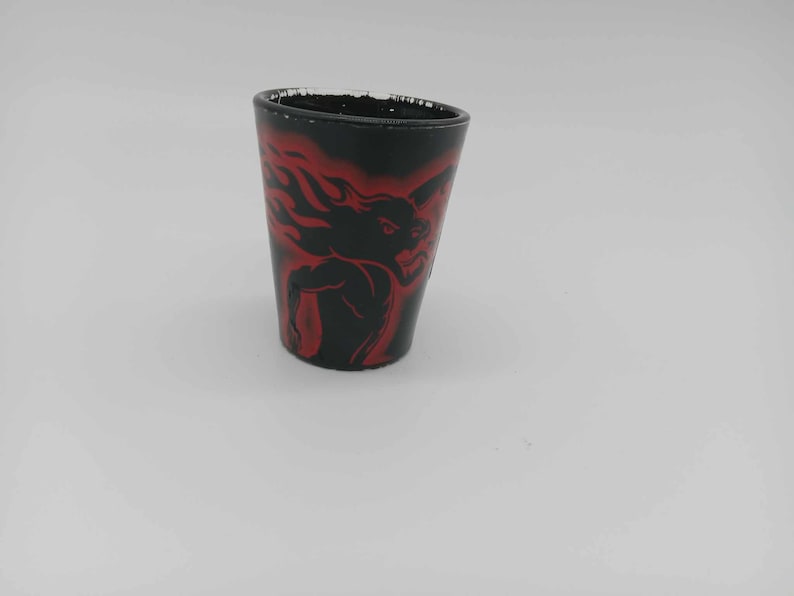 May include: Black shot glass with a red graphic of a muscular figure with flames. The shot glass has a slightly tapered shape and a black exterior with the red design. The rim of the glass has a distressed look.