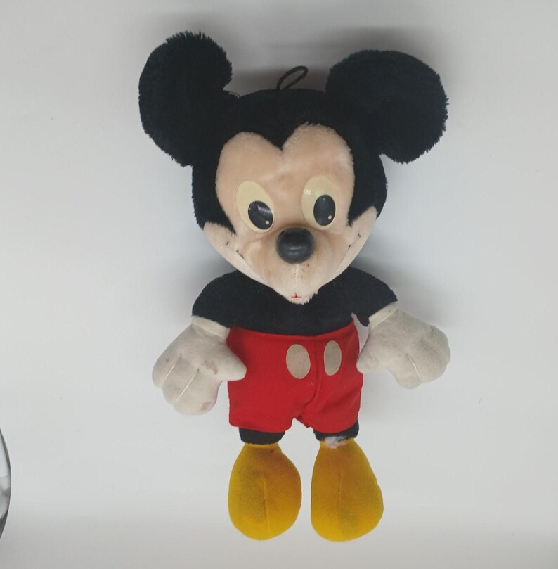 K&ouml;nnte beinhalten: Ein Mickey Mouse Pl&uuml;schpuppe mit schwarzen Ohren, einem schwarzen Hemd, roten Shorts, gelben Schuhen und wei&szlig;en Handschuhen. Die Puppe hat ein beiges Gesicht mit schwarzen Augen und einer schwarzen Nase. Die Puppe ist eine klassische Disney-Figur.