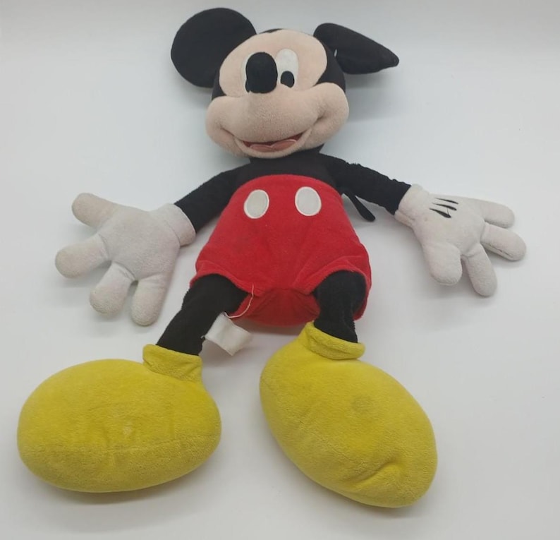 May include: A plush Mickey Mouse toy. The toy features a black head with large ears, a tan face, and a red shorts. The arms and legs are black, with white gloves and yellow shoes. The toy is a classic Disney character.