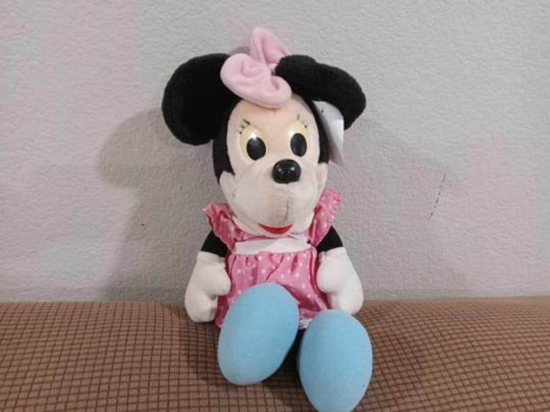 May include: A plush toy of Minnie Mouse wearing a pink bow and a pink and white polka dot dress. She has black ears and a white face with black eyes and a black nose. She is sitting with her legs crossed and her feet are blue.