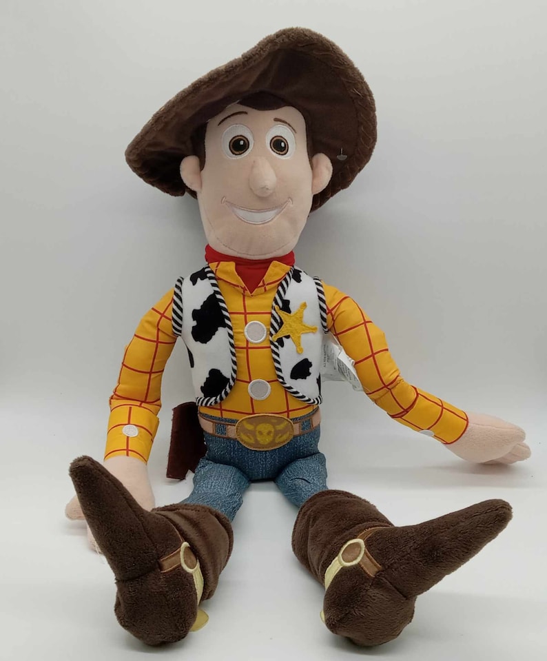 Puede incluir: Un peluche de Woody de Toy Story. Woody lleva un sombrero de vaquero marr&oacute;n, una camisa a cuadros amarilla, un chaleco blanco y negro, vaqueros azules y botas de vaquero marrones. Una estrella de sheriff dorada est&aacute; prendida en su chaleco.
