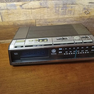 Puede incluir: Una radio reloj digital General Electric vintage con una pantalla fluorescente azul. La radio tiene una combinación de colores plateado y gris con botones para snooze, sleep y ajuste de pantalla. La radio también tiene control de volumen y banda.
