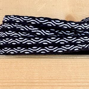 May include: Close-up of a woven black and white trim with a repeating wave pattern. The trim is layered and appears to be made of a soft material. The background is a light-colored wooden surface.
