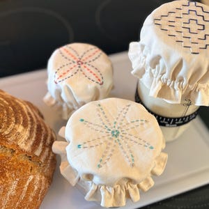 May include: A loaf of crusty bread next to three cream-colored jar covers with embroidered designs in red, blue, and teal. The covers have ruffled edges. The bread is golden brown.