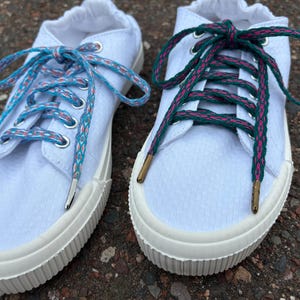 May include: Two white canvas sneakers with contrasting shoelaces. One pair features blue and coral patterned laces, the other has green and pink laces. The shoes have white rubber soles and silver and gold eyelets.