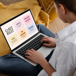 May include: A laptop displaying "Class Rules" with colorful boxes listing classroom guidelines. The laptop is open on a person's lap, with the person wearing a white shirt and blue jeans. A yellow pillow is in the background.