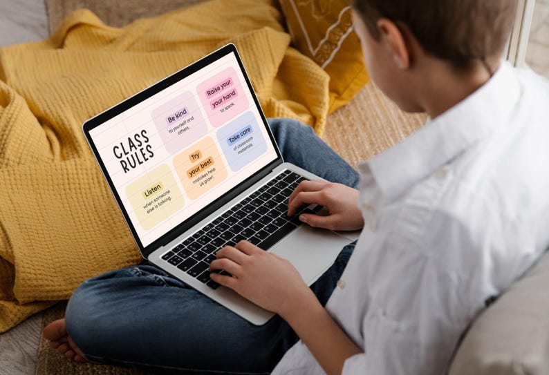 May include: A laptop displaying "Class Rules" with colorful boxes listing classroom guidelines. The laptop is open on a person's lap, with the person wearing a white shirt and blue jeans. A yellow pillow is in the background.