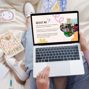 May include: A laptop displaying a presentation titled "About Me" with text and a photo of a classroom. Surrounding the laptop are children's drawings, including a drawing that says "I Love Mom". The person is wearing jeans and white shoes.