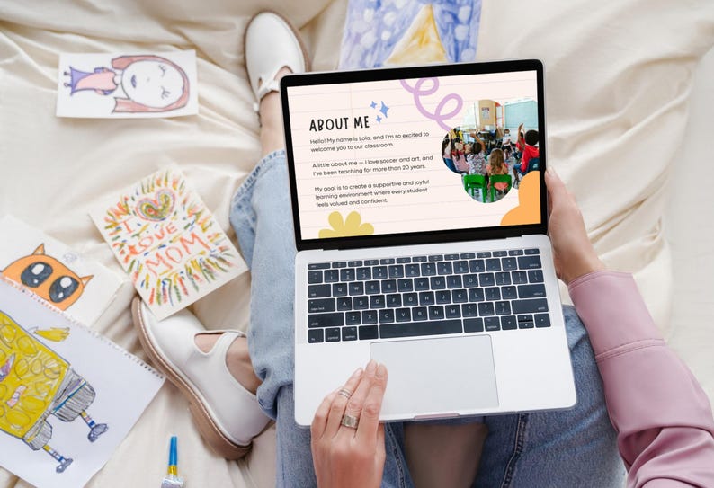 May include: A laptop displaying a presentation titled "About Me" with text and a photo of a classroom. Surrounding the laptop are children's drawings, including a drawing that says "I Love Mom". The person is wearing jeans and white shoes.