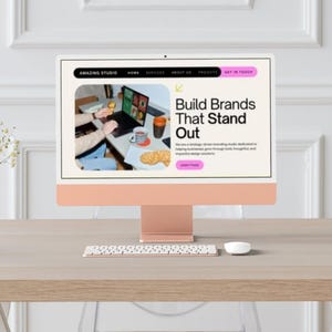 May include: A desktop computer with a light peach-colored stand and screen displays the text "Build Brands That Stand Out." A keyboard and mouse are in front of the screen. A vase of flowers sits to the left.