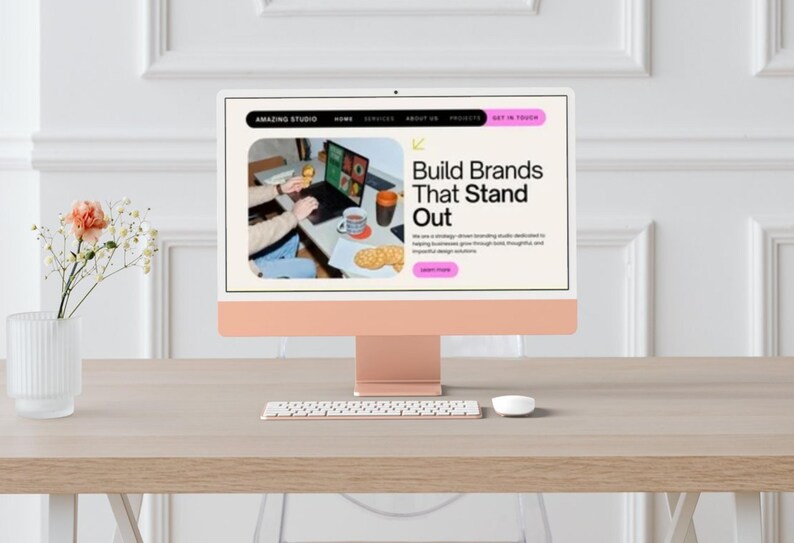 May include: A desktop computer with a light peach-colored stand and screen displays the text "Build Brands That Stand Out." A keyboard and mouse are in front of the screen. A vase of flowers sits to the left.