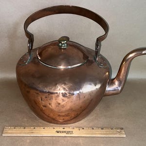 Puede incluir: Una tetera de cobre con un pico curvo y un asa grande. La tetera tiene una tapa con un pequeño pomo. Una regla de madera está en primer plano. La tetera mide aproximadamente 30 cm de altura.