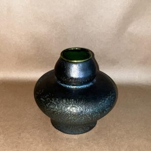 May include: A small, bulbous ceramic vase with a dark, metallic blue and black glaze featuring a textured, speckled finish. The vase has a narrow, rounded neck with a smooth, dark green interior rim and a wide, rounded body that tapers to a base.