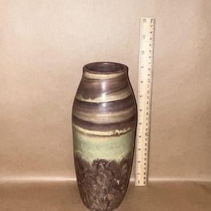 May include: A cylindrical ceramic vase with a tapered neck, featuring a dark brown base with textured patterns, a light green middle section, and a dark brown rim. A wooden ruler placed next to the vase shows a height of approximately 12 inches.