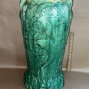 Puede incluir: Un jarrón alto de cerámica verde con un diseño en relieve detallado de peces, plantas acuáticas y flores. El jarrón tiene un borde ancho y festoneado y una superficie texturizada. Una regla está al lado.