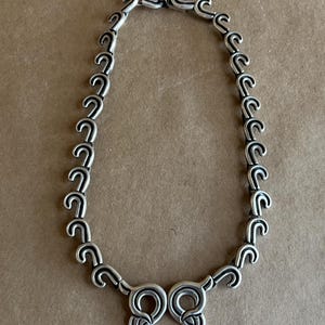 Op de afbeelding: Een zilverkleurige ketting met een herhalend, gestileerd haakontwerp. De ketting bestaat uit onderling verbonden gebogen elementen, waardoor een doorlopend patroon ontstaat. De sluiting heeft een decoratief, symmetrisch ontwerp. De ketting wordt getoond tegen een neutrale achtergrond.