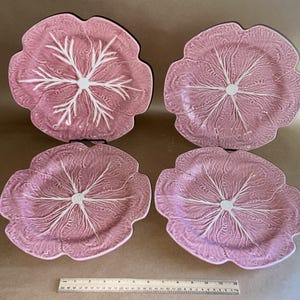 May include: Four pink ceramic plates shaped like cabbage leaves with white veining radiating from the center. A wooden ruler is placed horizontally at the bottom, showing the plates measure approximately 12 inches across at their widest point.
