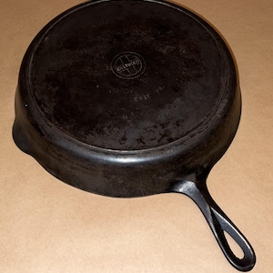May include: A vintage Griswold cast iron skillet, viewed from the bottom. The dark, well-seasoned pan features a long handle with a hole for hanging. The text "Griswold Erie PA" is embossed on the base, indicating its origin.
