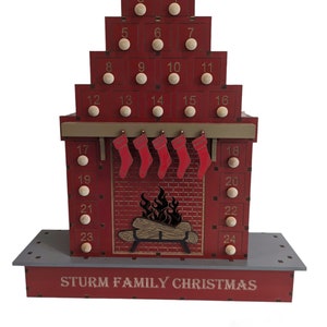 May include: A red wooden Christmas advent calendar shaped like a house with a chimney and a fireplace. The calendar has 24 numbered doors, each with a small wooden knob. The doors are decorated with gold numbers and the text "STURM FAMILY CHRISTMAS".