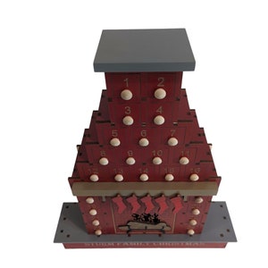 May include: A red wooden advent calendar shaped like a house with a chimney and a fireplace. The calendar has 16 numbered doors, each with a small wooden knob. The fireplace has stockings hanging from it. The text "STORM FAMILY CHRISTMAS" is printed on the base of the calendar.