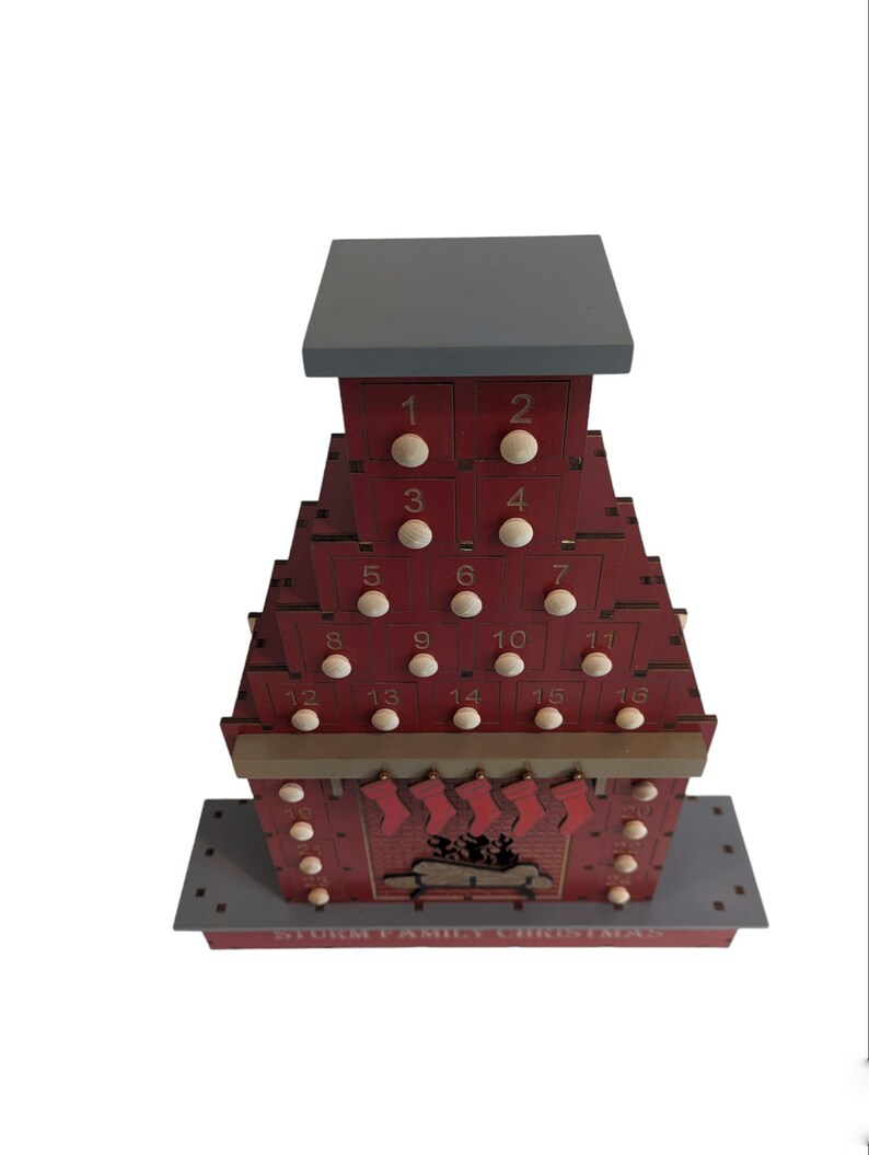 May include: A red wooden advent calendar shaped like a house with a chimney and a fireplace. The calendar has 16 numbered doors, each with a small wooden knob. The fireplace has stockings hanging from it. The text "STORM FAMILY CHRISTMAS" is printed on the base of the calendar.