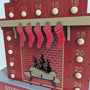May include: A red wooden advent calendar shaped like a fireplace with 24 numbered doors. The doors are decorated with wooden circles. There are five stockings hanging from the mantel with names written on them: Ryan, Leanne, Ryleigh, Coltan, and Raegan. The fireplace has a brick design and a fire burning inside. The bottom of the calendar says "STURM FAMILY CHRISTMAS".