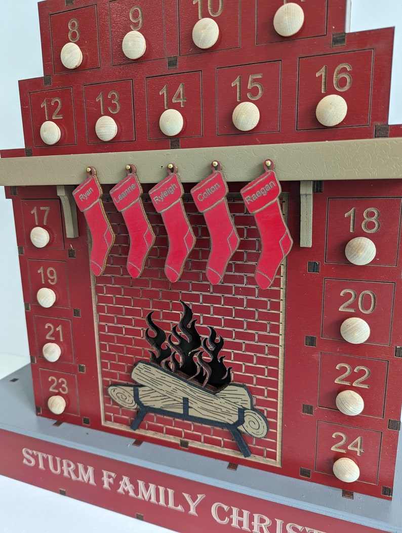 May include: A red wooden advent calendar shaped like a fireplace with 24 numbered doors. The doors are decorated with wooden circles. There are five stockings hanging from the mantel with names written on them: Ryan, Leanne, Ryleigh, Coltan, and Raegan. The fireplace has a brick design and a fire burning inside. The bottom of the calendar says "STURM FAMILY CHRISTMAS".