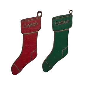 May include: Two wooden Christmas stocking ornaments with the name "Colton" written on them. One stocking is red and the other is green.