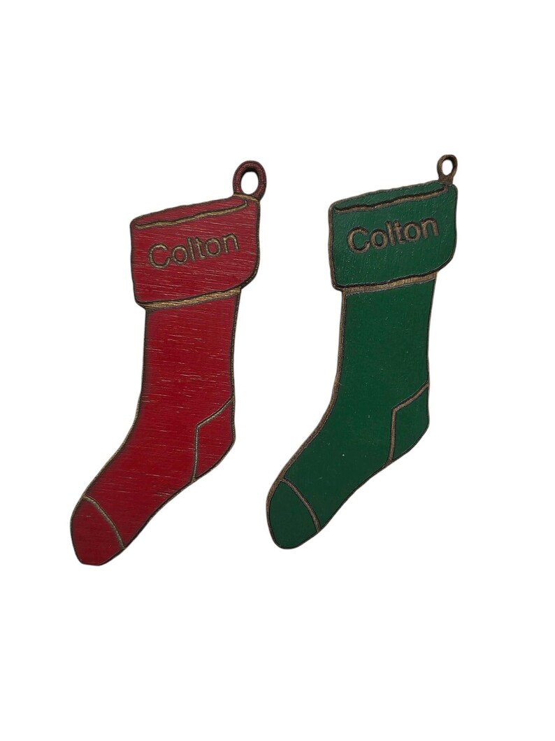 May include: Two wooden Christmas stocking ornaments with the name "Colton" written on them. One stocking is red and the other is green.