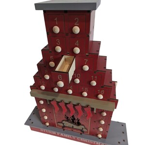 May include: A red wooden Christmas advent calendar shaped like a fireplace with a chimney and stockings. The calendar has 24 numbered drawers, each with a small white knob. The base of the calendar reads "STORM FAMILY CHRISTMAS".