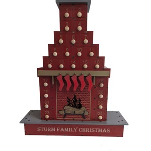 May include: A red wooden advent calendar shaped like a fireplace with a chimney on top. The calendar has 24 numbered doors, each with a small white knob. The fireplace has a brick pattern and a black cutout of a fire inside. The calendar is decorated with red stockings and the text "STURM FAMILY CHRISTMAS".