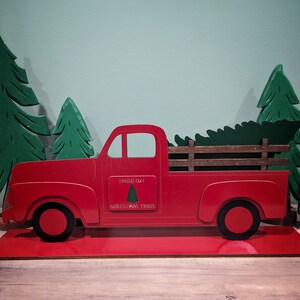 May include: A red wooden pickup truck carrying a green Christmas tree, with the words "Fresh Cut Christmas Trees" on the side. The truck is flanked by green wooden Christmas trees, all set on a red base. A festive holiday decoration.