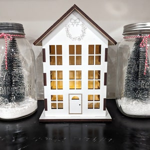 May include: A white wooden house-shaped decoration with multiple windows and a door, flanked by two glass mason jars. Each jar contains a small Christmas tree and artificial snow, and is adorned with a red and white twine bow. The house has a wreath design.